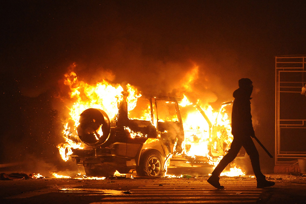 Silhouette of figure holding a baseball bat and walking away from car on fire.