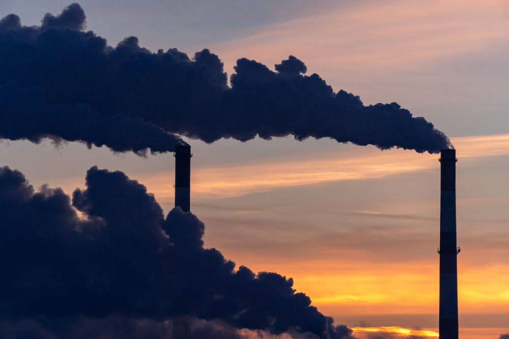 Silhouettes of smoky emissions from three smokestacks at sunset.