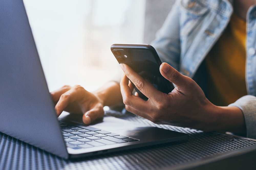 A person working on a laptop while holding a cell phone in one hand