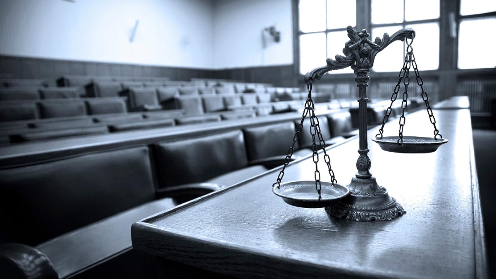 The scales of justice sit on a table in front of an empty jury box in a court room.