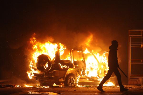 A silhouetted figure carrying a baton walks away from a burning car.